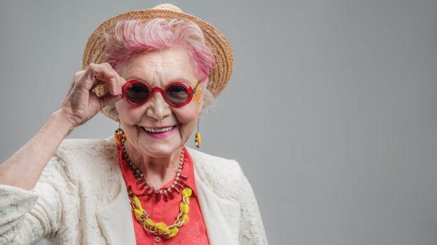 kolorowo ubrana uśmiechnięta starsza kobieta podtrzymuje dłonią okulary i patrzy w aparat