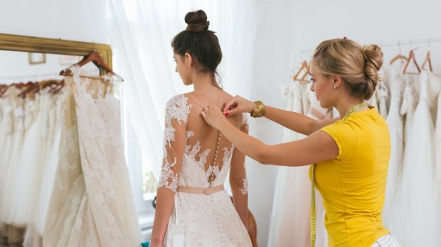 dressmaker helping the bride to put her wedding dress on in clothes shop.