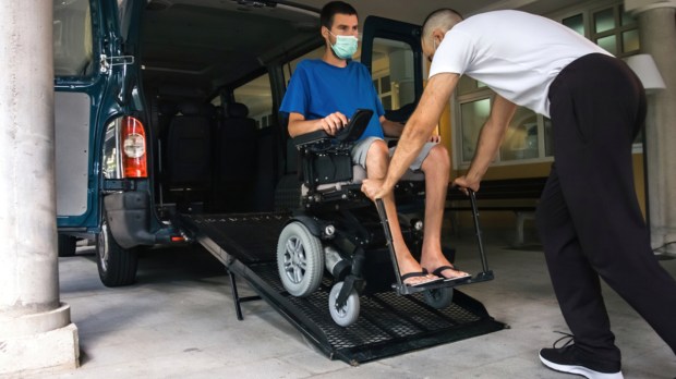 Man on wheelchair using accessible vehicle with ramp for transportation with driver helping.