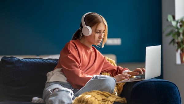 Focused teenage girl wearing headphones typing text on laptop relaxed sitting on couch in living room